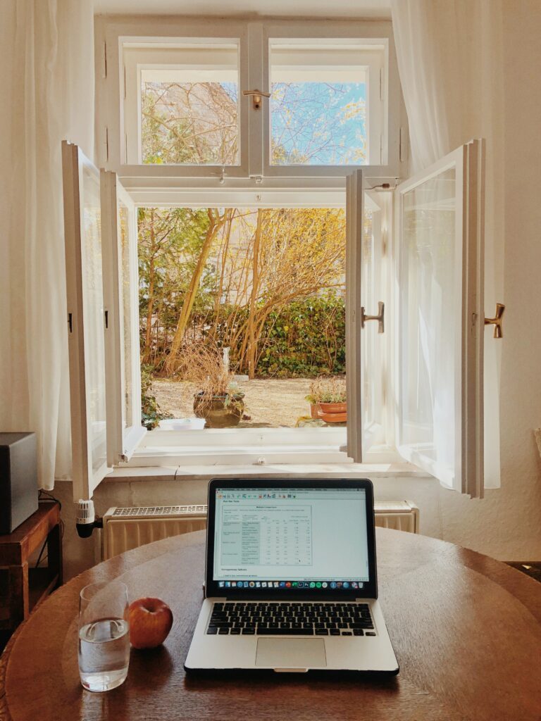 une fenêtre ouverte sur un jardin. Au premier plan, une table en bois sur lequel se trouve un ordinateur portable