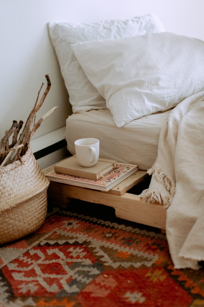 image d'un lit avec des draps beiges, 2 oreillers blancs, une table de chevet en bois blond sur lequel sont posés des livres et une tasse en céramique.