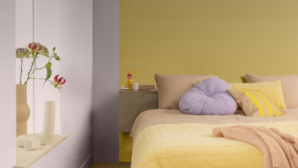 cette photo représente une chambre, et plus particulièrement le lit et la tête de lit. La tête de lit est peinte en jaune moutarde, le mur a coté est en rose pastel