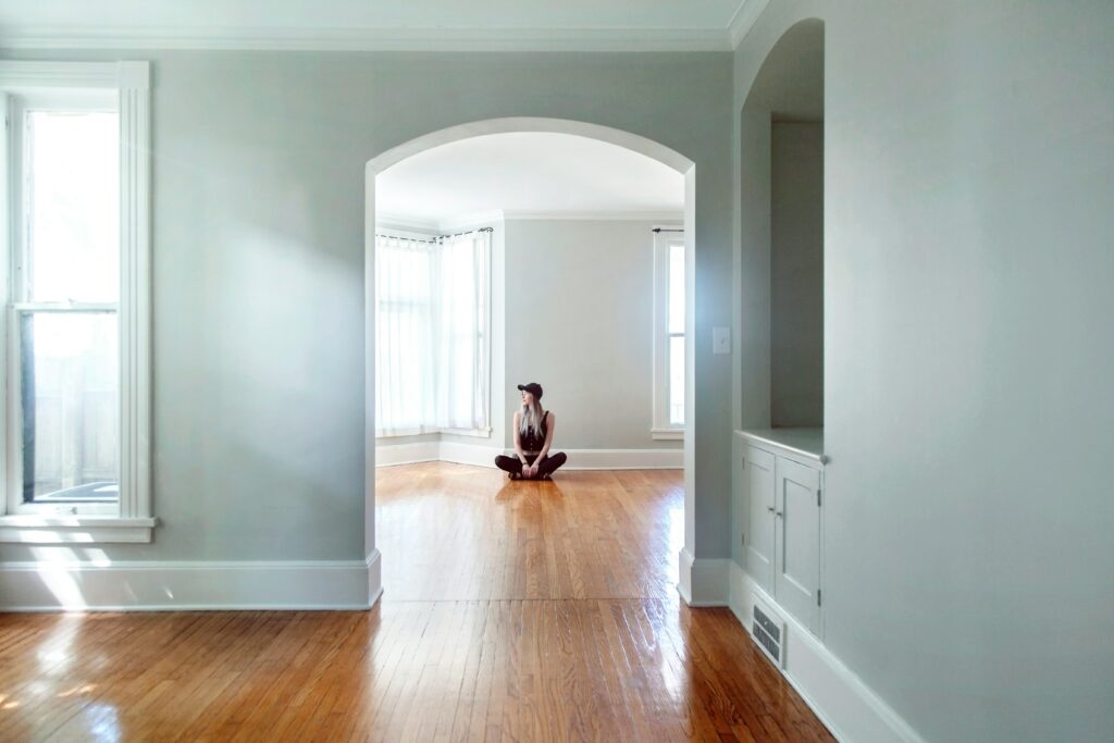 Jeune femme assise en tailleur dans un grand salon vide et lumineux, au centre d'une arche architecturale, illustrant l'introspection et la recherche d'harmonie intérieure en psychologie de l'habitat.