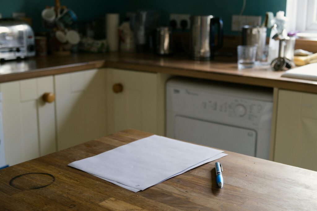 Feuilles de papier blanches et stylo posés sur une table en bois avec une trace de tasse, illustrant le manque d'espace et le mal-être chez soi en télétravail.