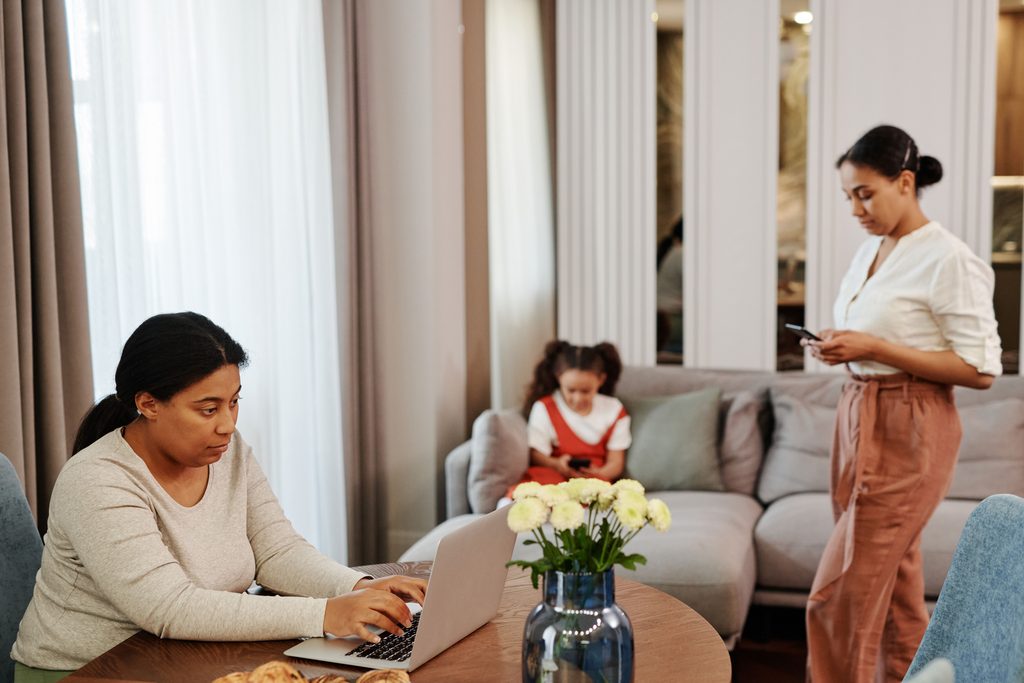 Une maman travaillant sur ordinateur portable à la maison tandis que sa fille ado est sur son téléphone et la plus jeune sur une tablette.